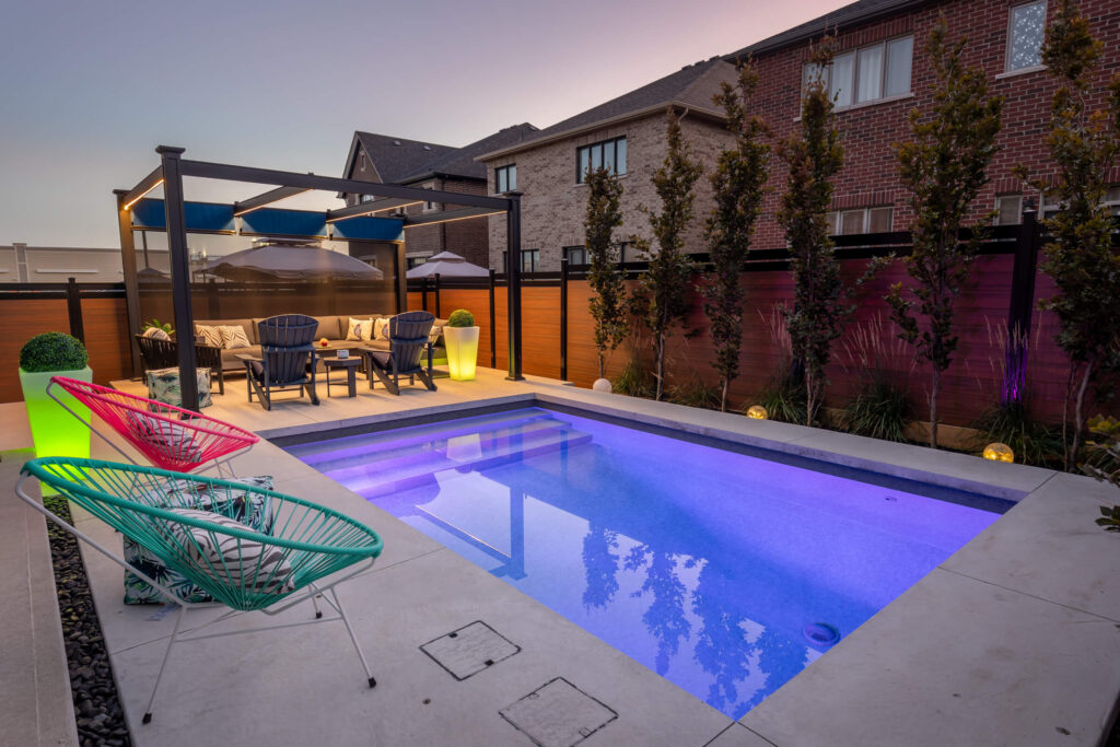 A modern backyard with a lit swimming pool at dusk, featuring colorful chairs, a seating area under a pergola, and ambient lighting. Brick houses and a fence surround the space.