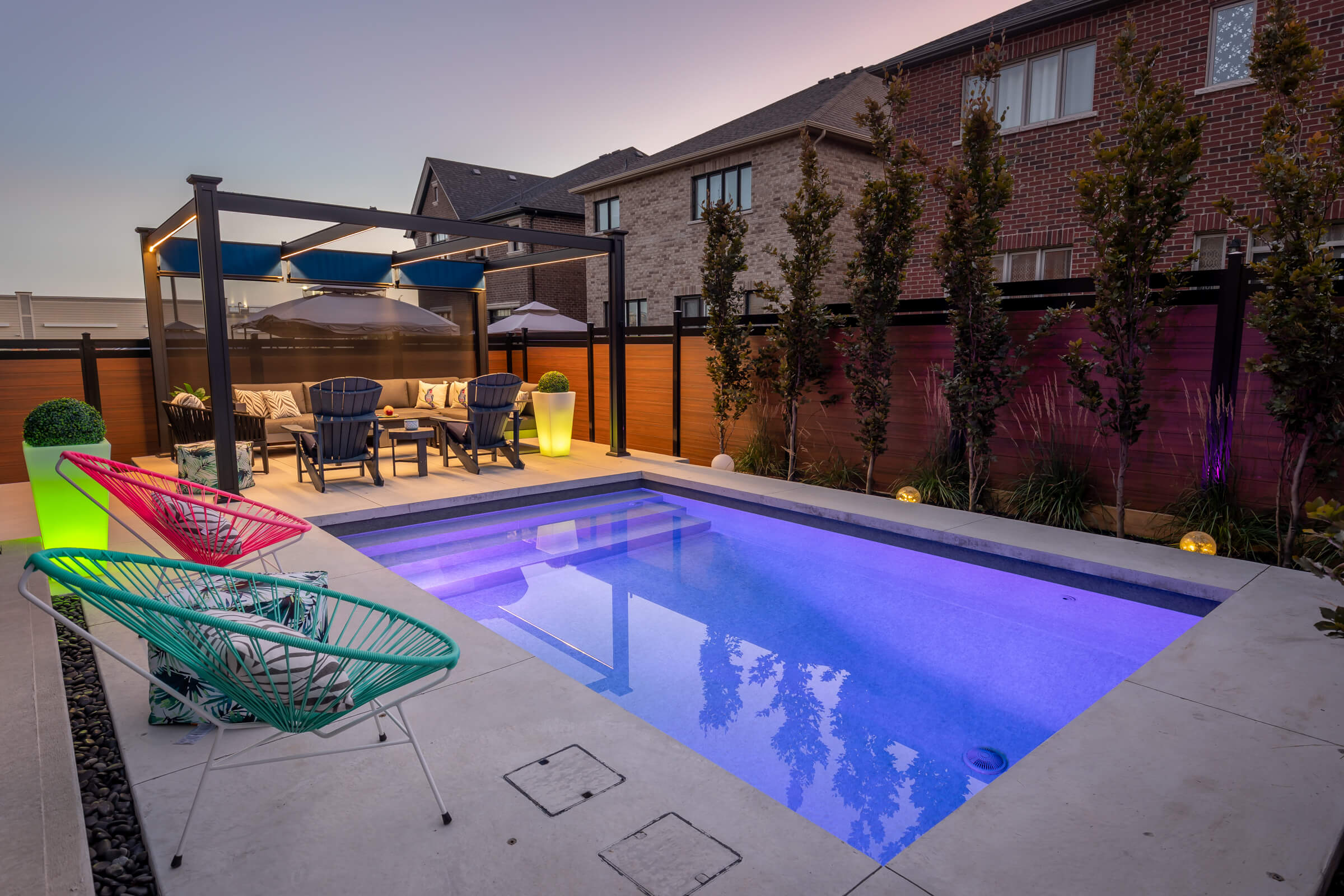 A modern backyard with a lit swimming pool at dusk, featuring colorful chairs, a seating area under a pergola, and ambient lighting. Brick houses and a fence surround the space.