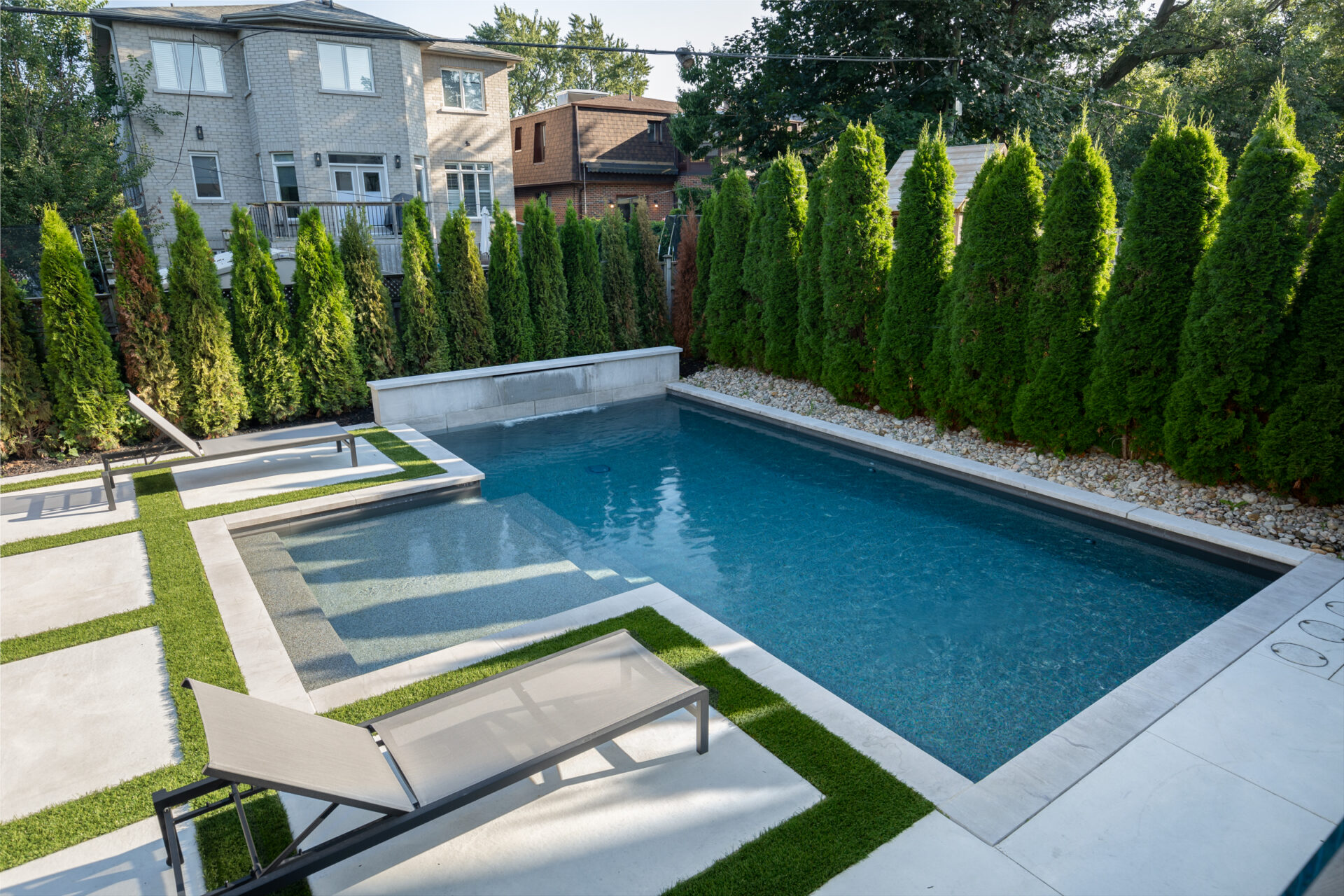 This image shows a modern backyard with a rectangular swimming pool, lounge chair, artificial grass, stepping stones, and tall privacy hedges.