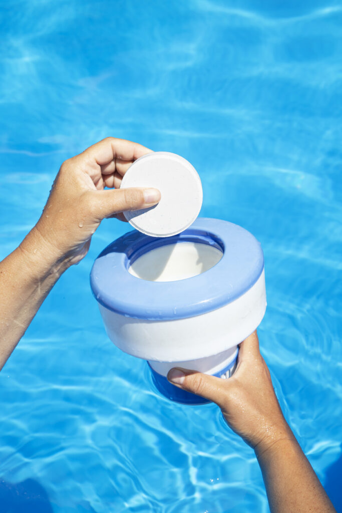 A person's hands are holding a floating chlorine dispenser in a swimming pool, the lid being opened to reveal the compartment inside.