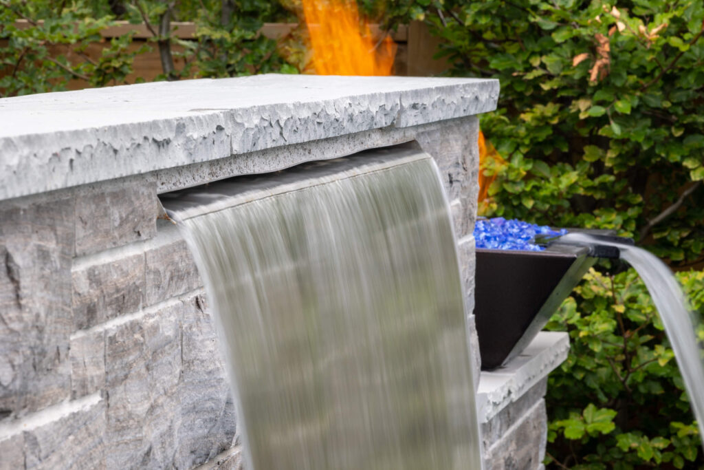 An outdoor stone fireplace with a smooth water feature spilling out. A fire crackles in the background amidst lush green foliage.