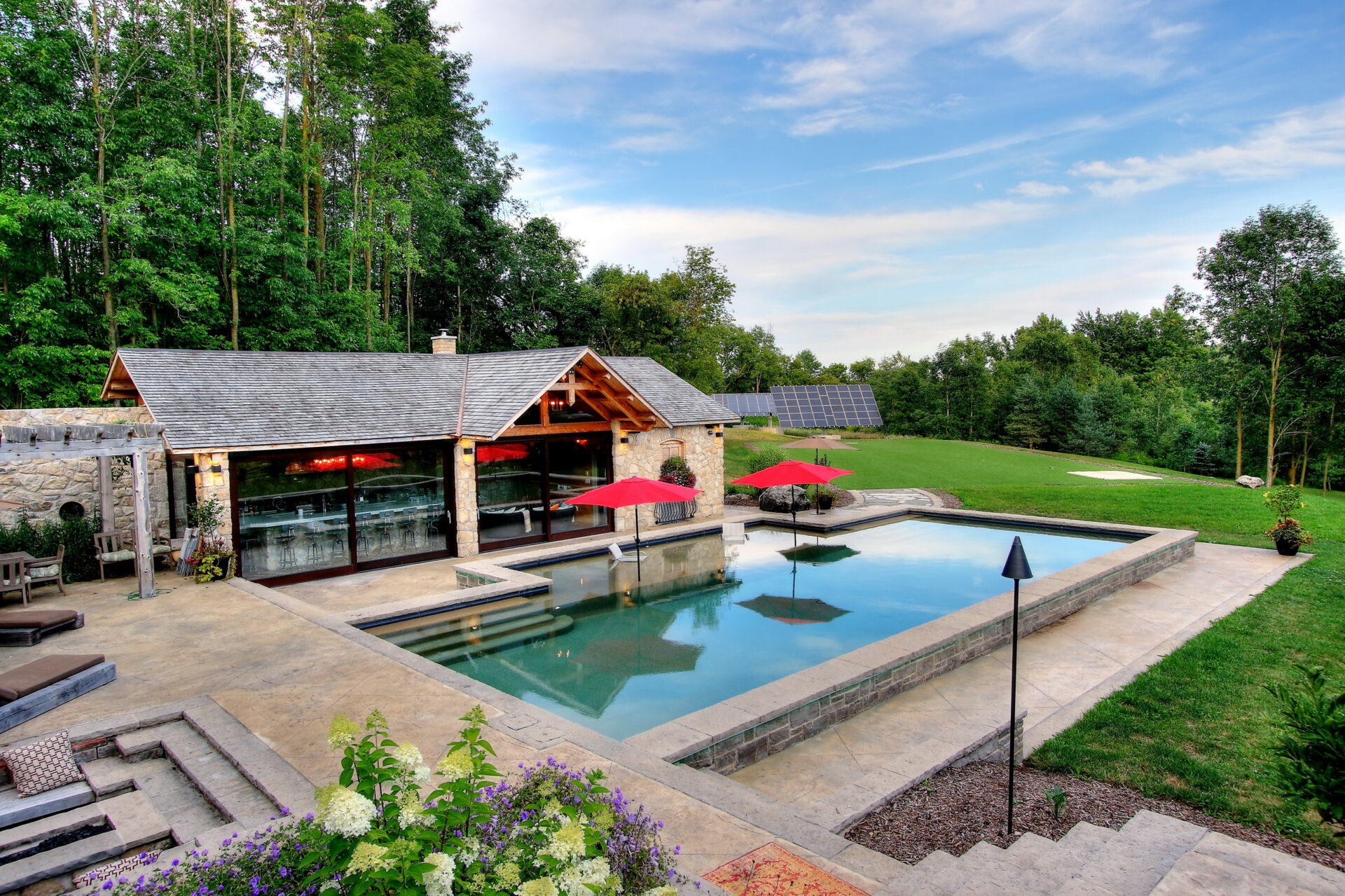 An upscale backyard with a rectangular pool, lounging chairs, red umbrellas, stone house, lush trees, clear sky, and solar panels in the distance.