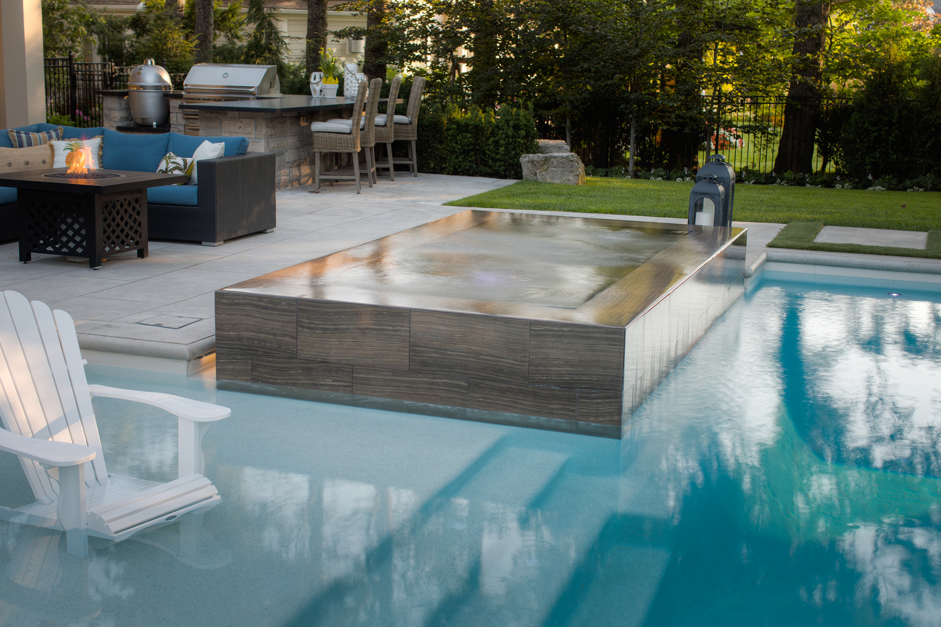 A luxurious backyard with an in-ground pool, hot tub, outdoor seating, fireplace, and BBQ grill. A white Adirondack chair is by the poolside.
