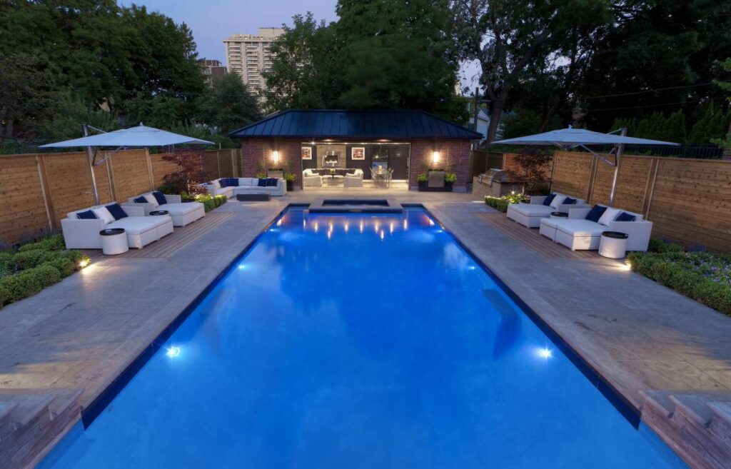 An elegant outdoor pool area during twilight with lounge furniture, umbrellas, and ambient lighting—adjacent to a cozy living space and wooden fence.