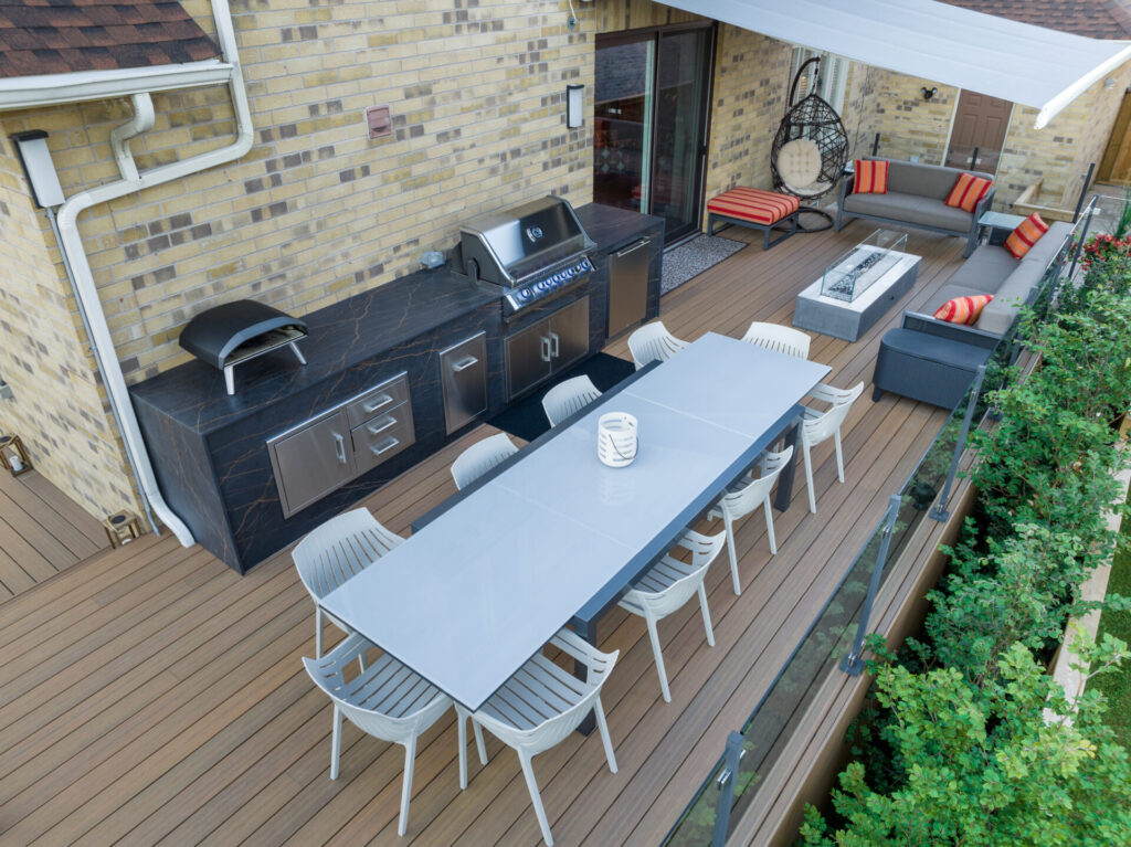 An outdoor patio featuring a modern kitchen setup with a grill, a dining table with chairs, and a lounge area with a sofa and armchair.