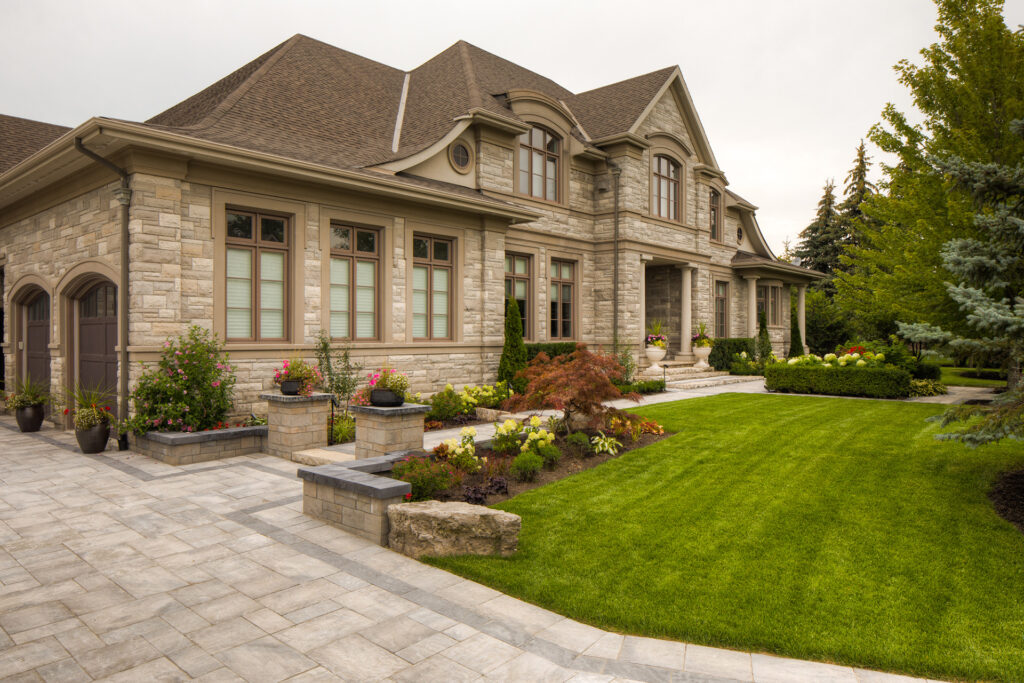 Large elegant stone house with manicured lawn, landscaped garden, and paved walkway, featuring multiple windows, a peaked roof, and ornamental plants.