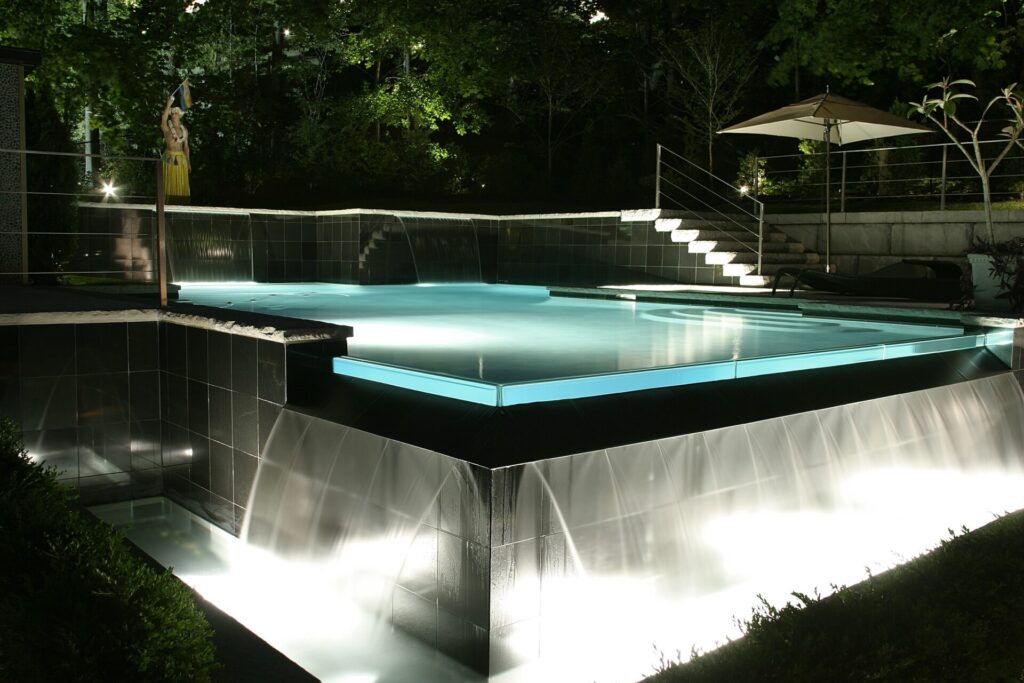 An illuminated outdoor swimming pool at night, with cascading water features and steps leading into the water, surrounded by trees and an umbrella.