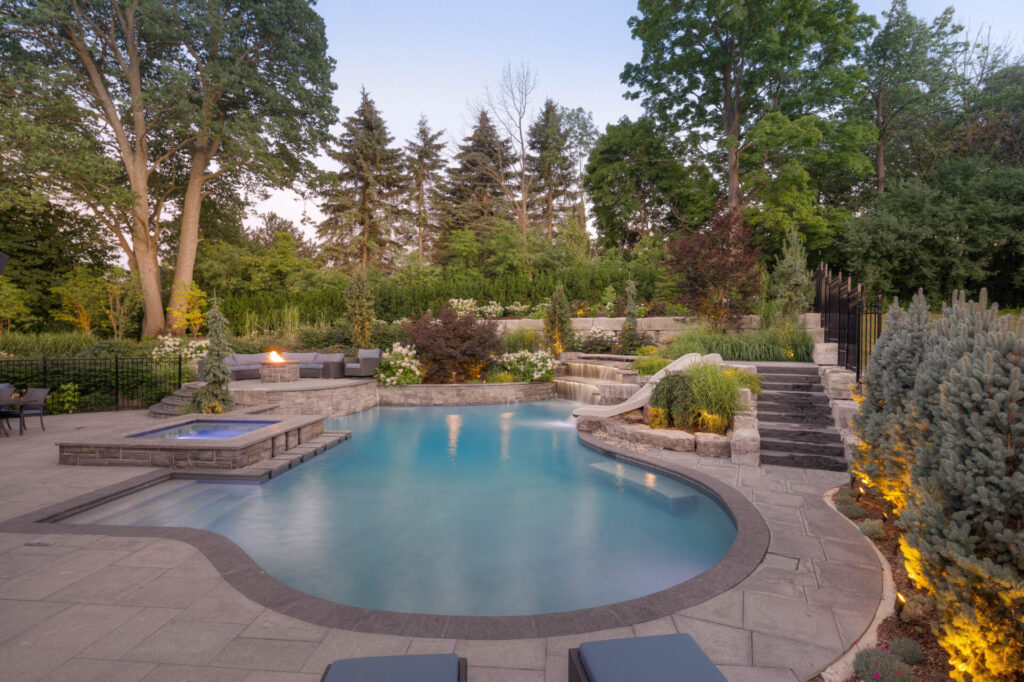 An elegant backyard with a curvy swimming pool at dusk, surrounded by well-manicured gardens, stone steps, a fire feature, loungers, and tall trees.