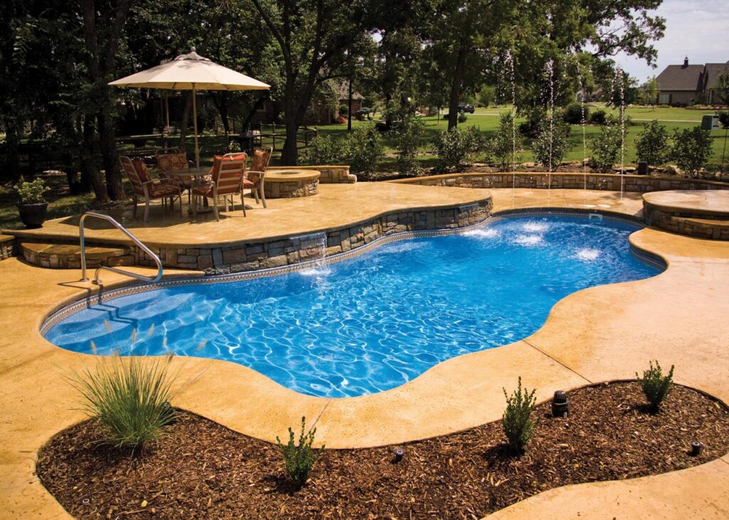 A kidney-shaped swimming pool with clear blue water, surrounded by a stone deck and landscaped garden, featuring patio furniture and a parasol.