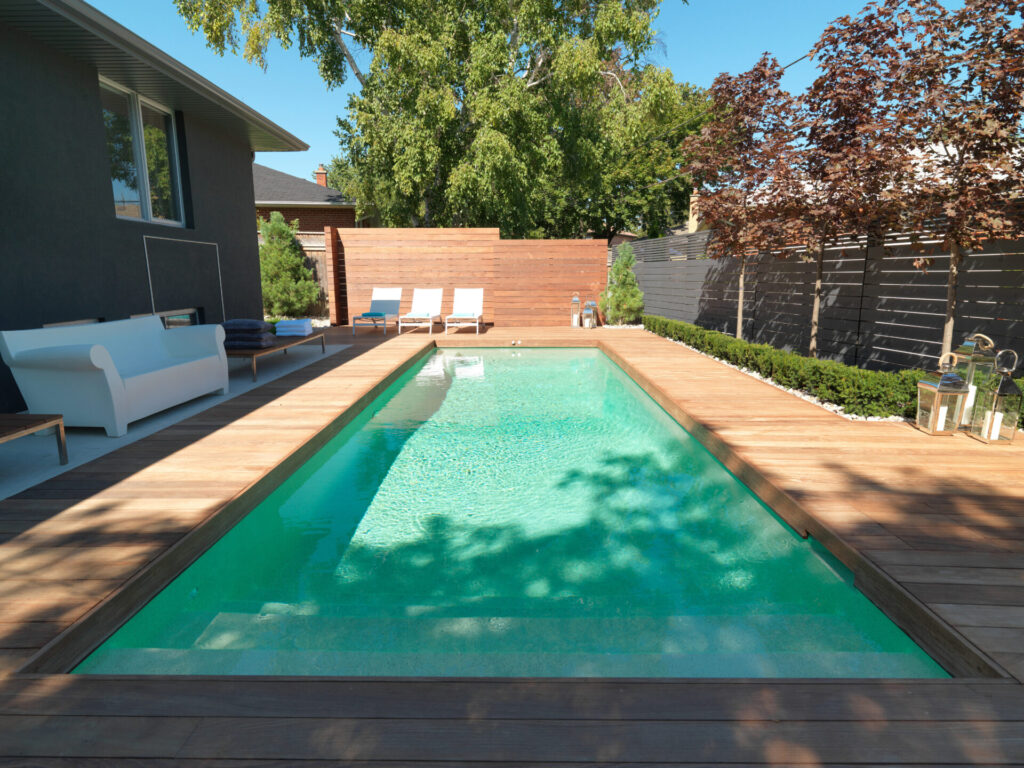 A modern backyard with a rectangular swimming pool, wooden deck, stylish outdoor furniture, trees, and a high privacy fence under a clear blue sky.