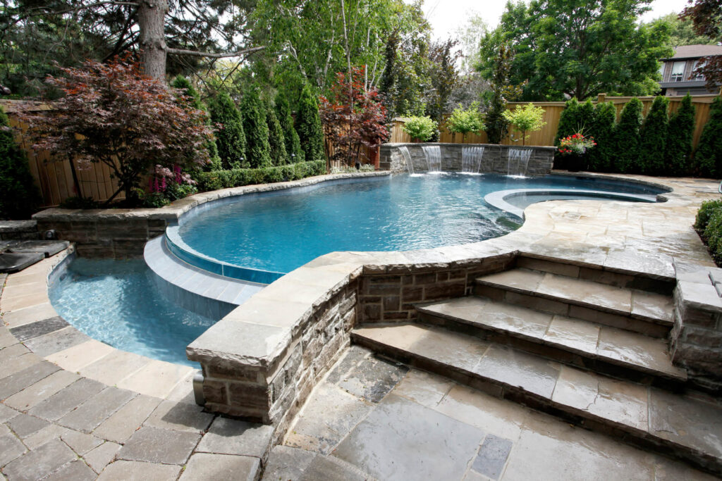 An elegant backyard with a kidney-shaped pool, stone steps, a curved water feature, lush trees, and a neatly manicured lawn under a cloudy sky.