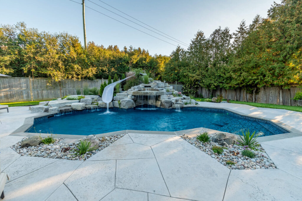 A luxurious backyard swimming pool with a water slide and rock feature, surrounded by a tiled patio, landscaping, and a wooden fence, under a clear sky.