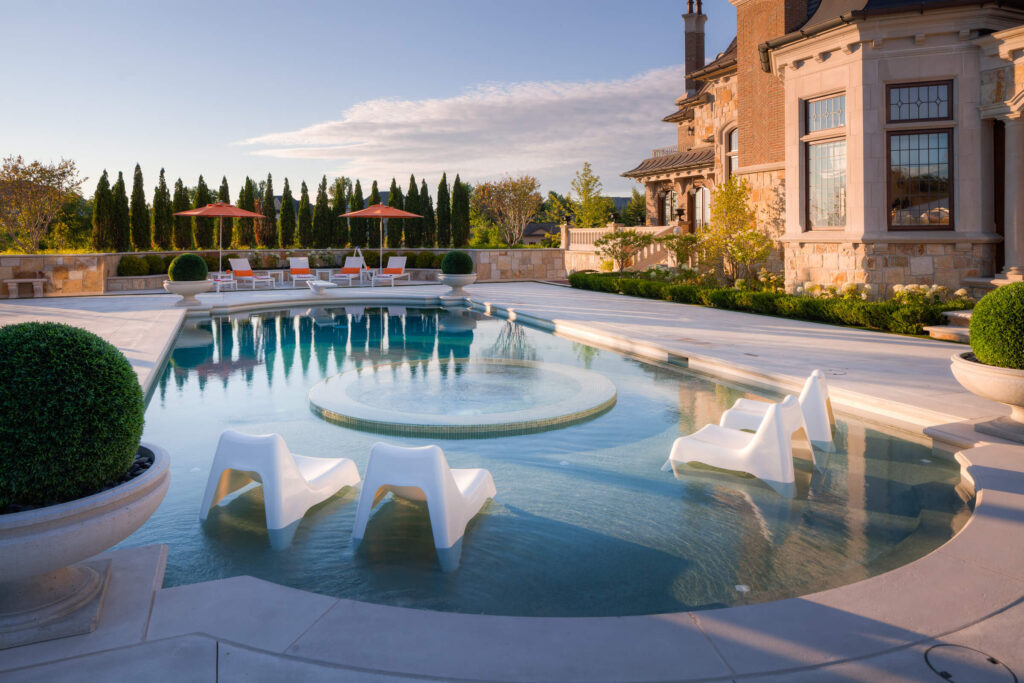 This image shows a luxurious outdoor swimming pool with sun loungers, parasols, surrounded by topiary and a stately home in the background.