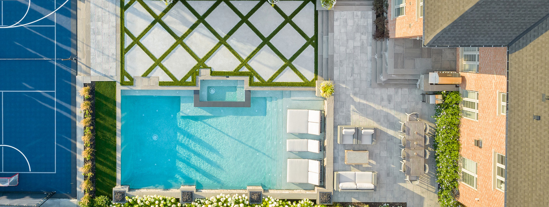 Aerial view of a luxurious backyard with a large swimming pool, an adjacent tennis court, lounging chairs, manicured hedges, and a part of a house.