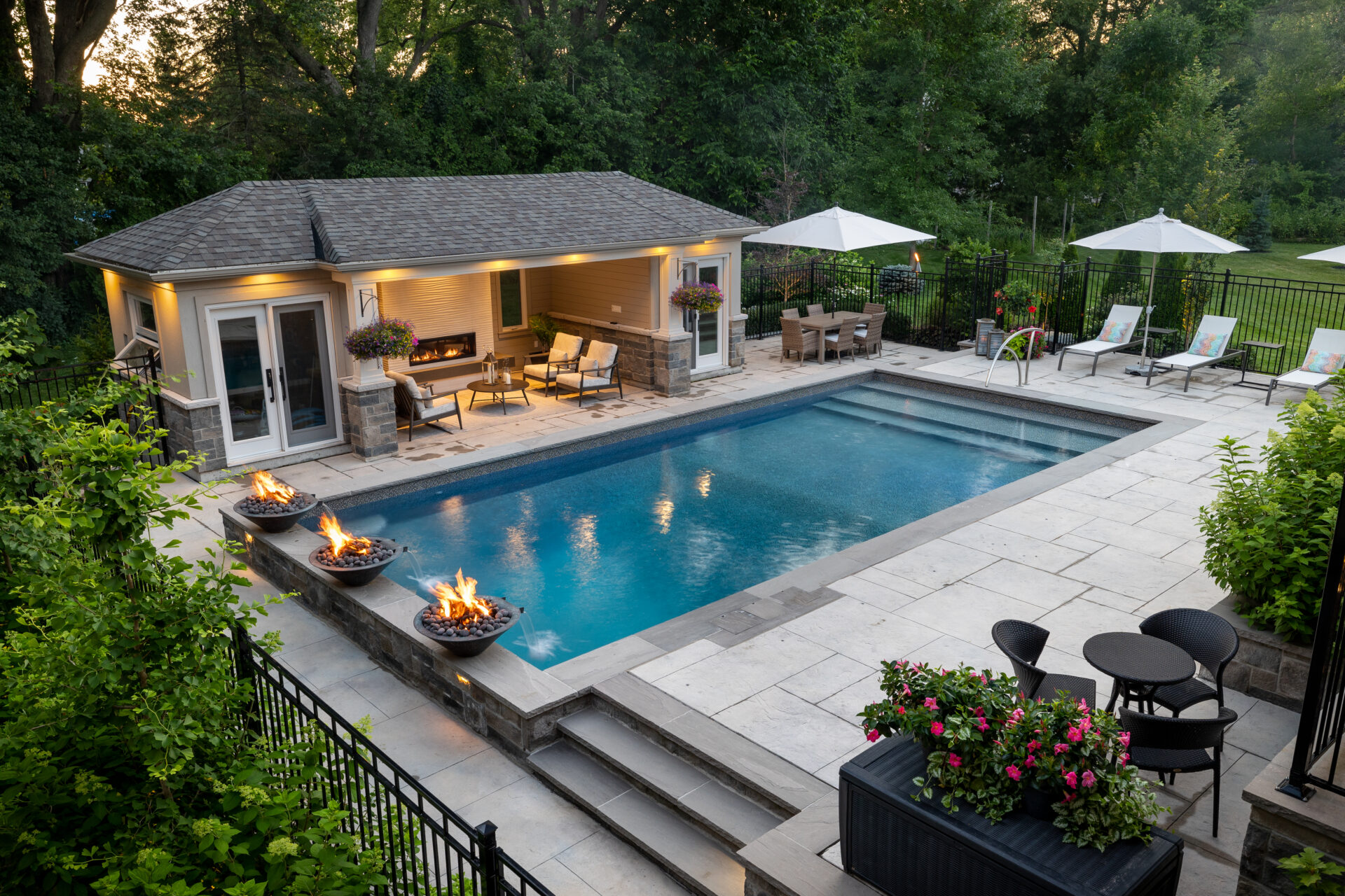 An upscale backyard features a pool, patio with seating and dining areas, umbrellas, greenery, and lit fire bowls at dusk.