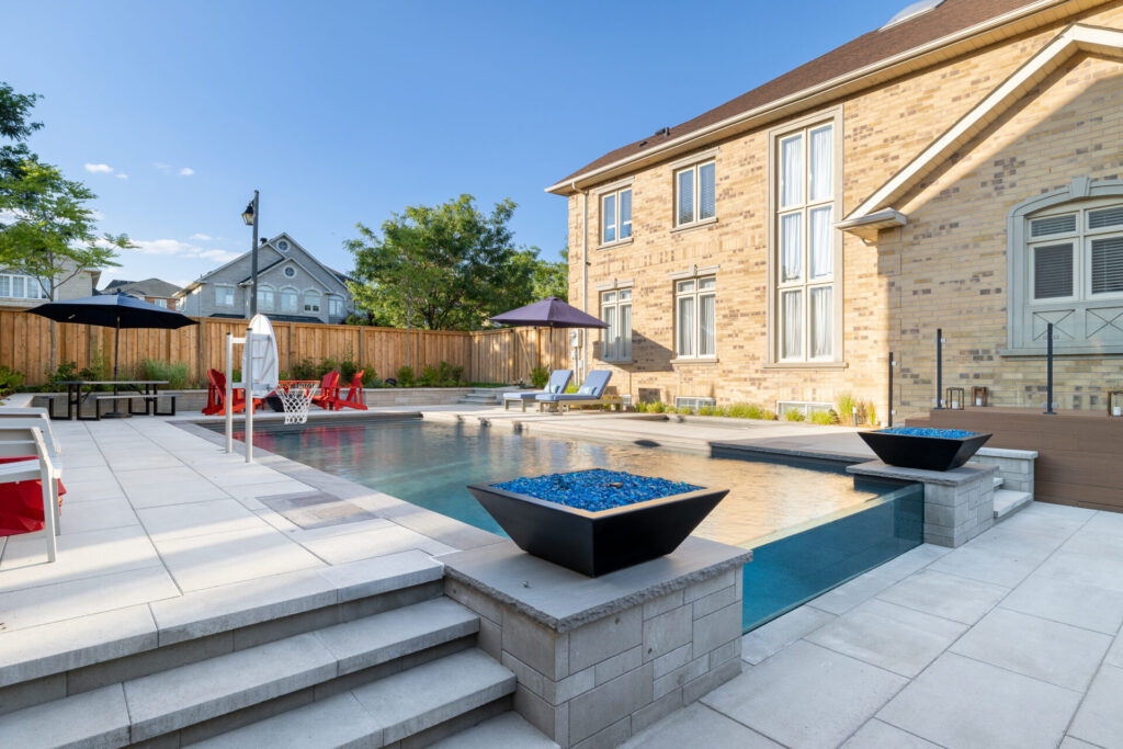 A luxurious backyard with a swimming pool, lounging chairs, umbrellas, and a fire pit. Stone patio and steps lead to a well-maintained house with large windows.