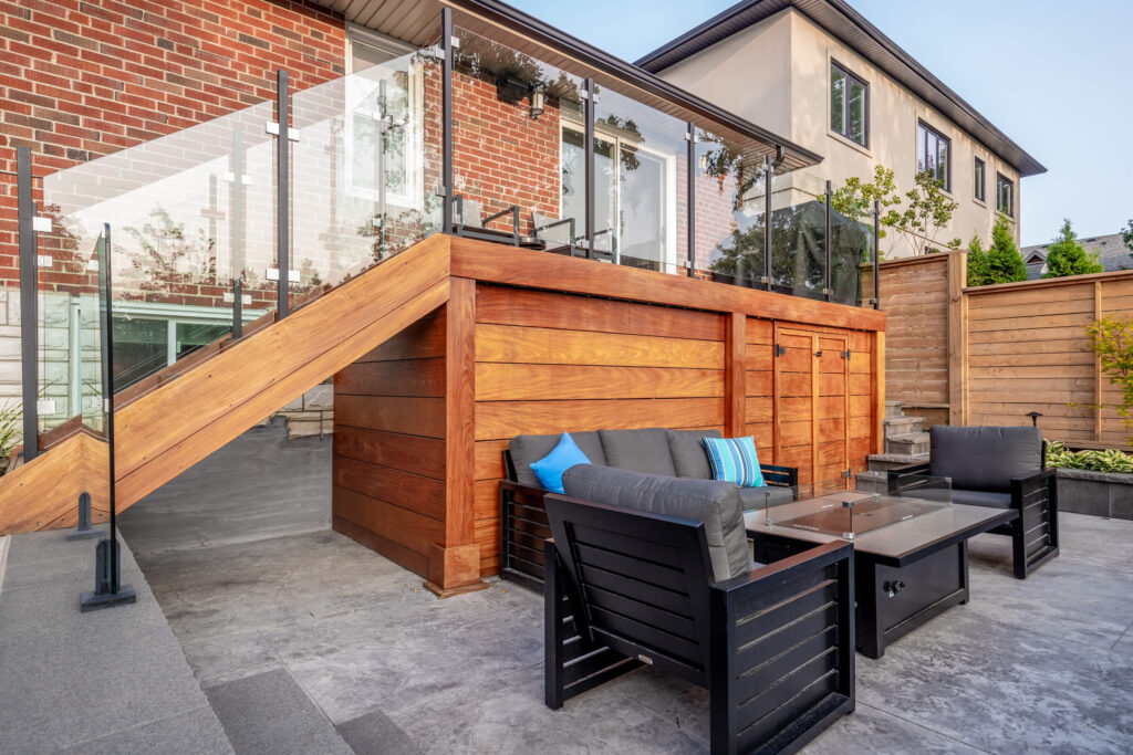 Modern outdoor patio with wooden staircase, glass railing, stylish furniture, and brick wall. Cozy space with cushions, plants, and a clear sky.