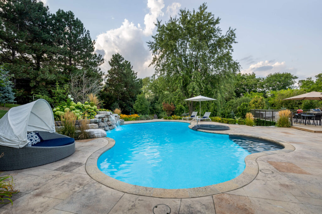A kidney-shaped swimming pool is surrounded by lush landscaping, lounge chairs, a daybed, and a patio area, creating a serene backyard oasis.