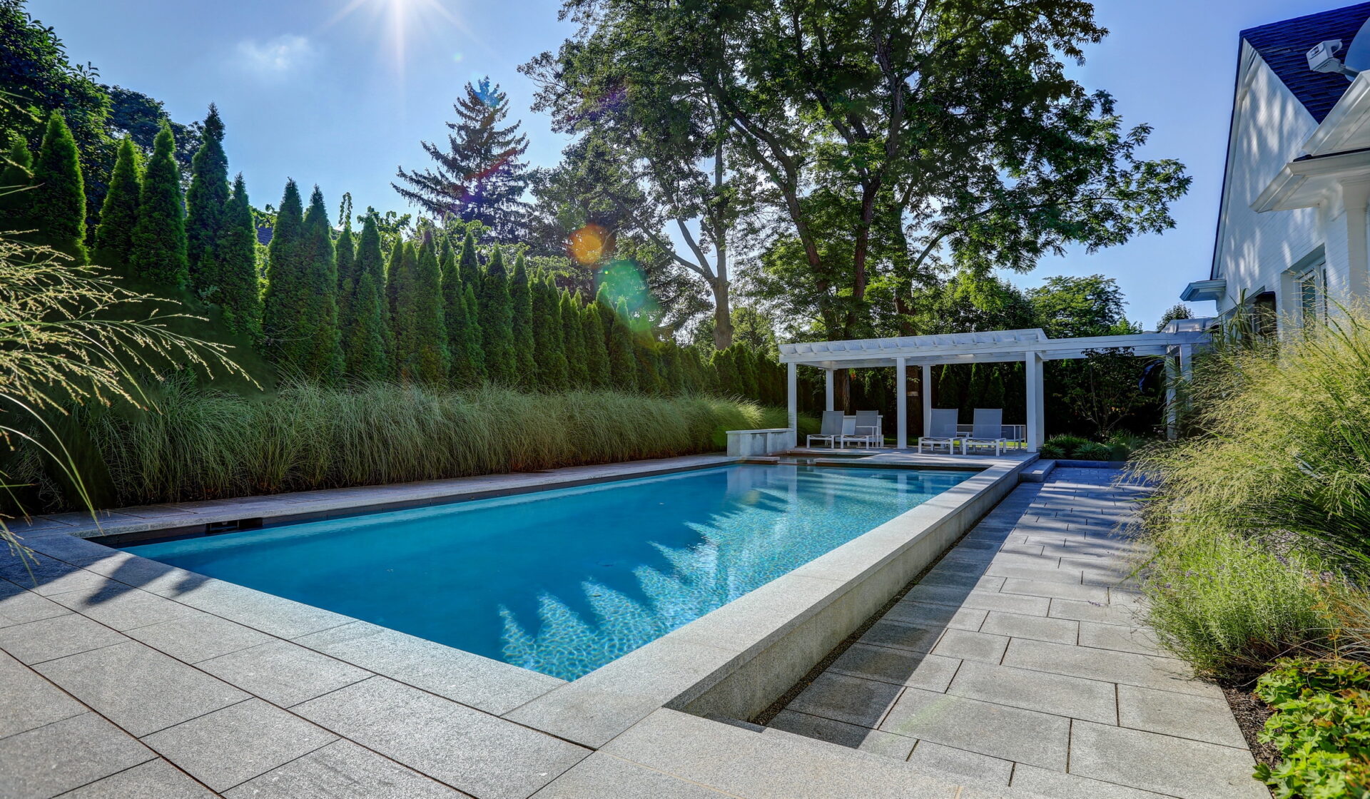 A serene backyard featuring a rectangular swimming pool, a white pergola with seating, lush greenery, and a blue sky peeking through the trees.