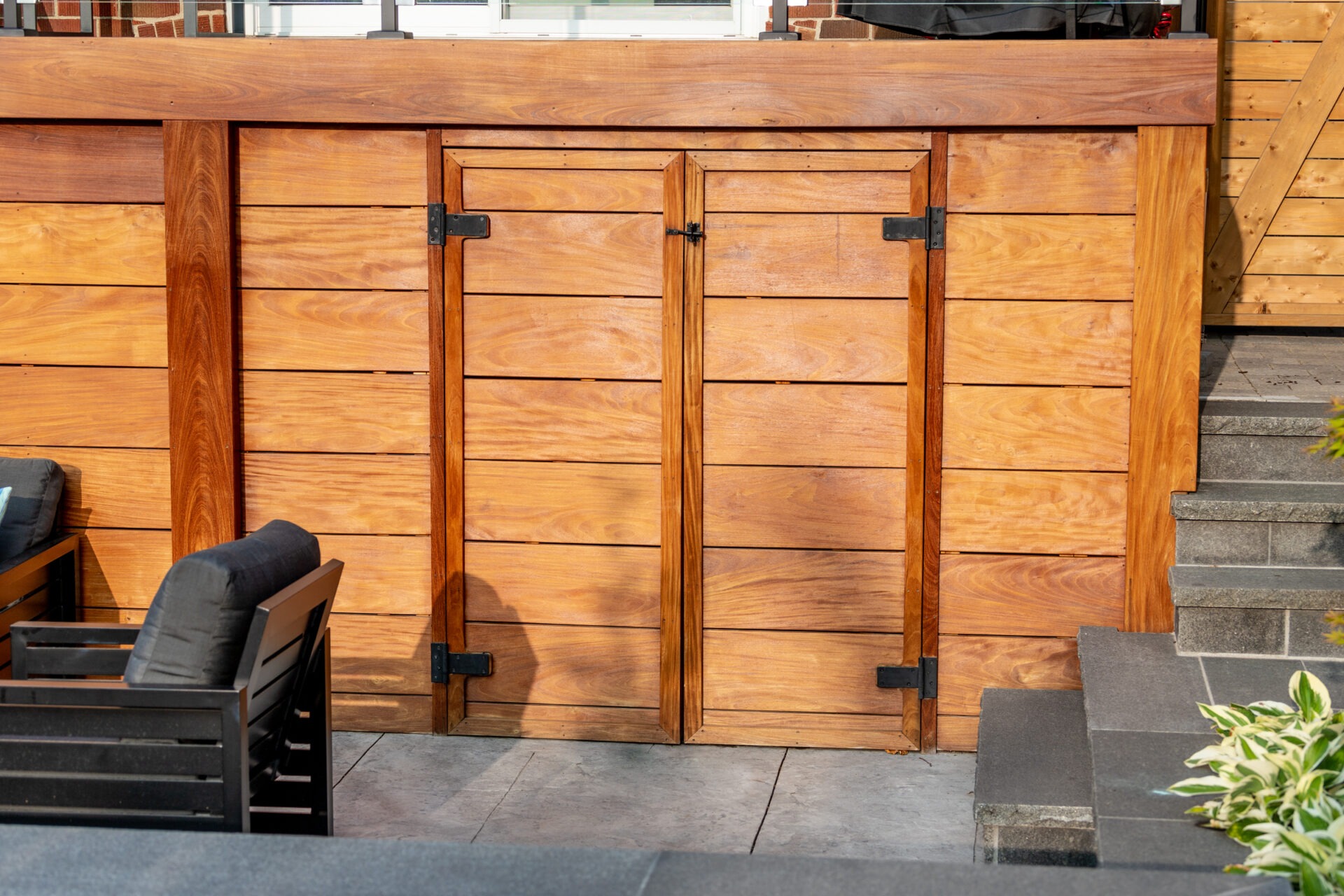 This image shows a set of wooden gates in a fence, with metal hardware, adjacent to stairs and a deck with outdoor furniture and plants.