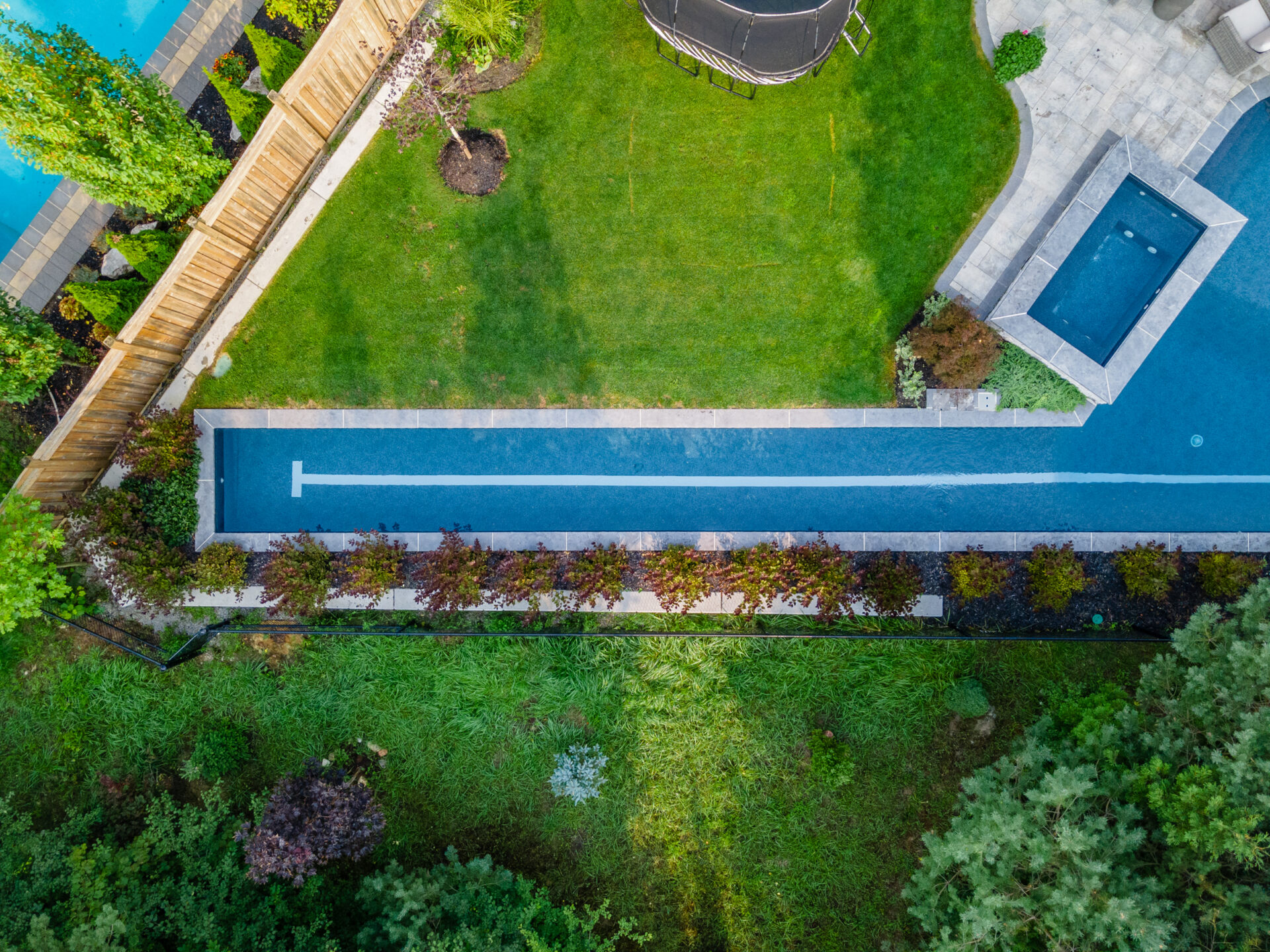 An aerial view of a well-manicured backyard with a long, narrow swimming pool, green lawn, landscaping, a hot tub, and a trampoline.