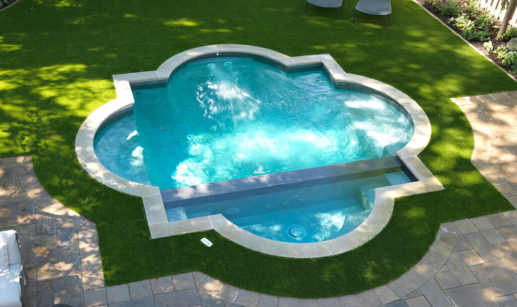 This image shows a uniquely shaped swimming pool surrounded by artificial grass and stone paving, with sunlight casting dappled shadows from nearby trees.