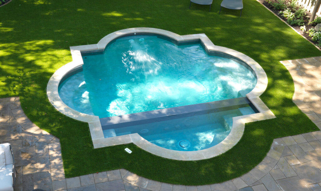 An aerial view of a vibrant blue, uniquely shaped swimming pool set in green artificial turf with surrounding patio and lounge chairs, on a sunny day.