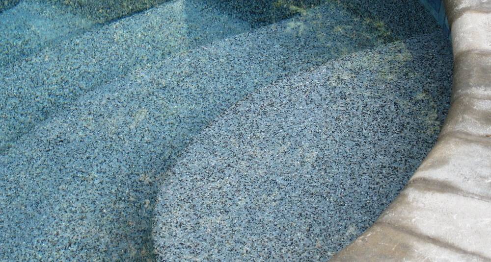 The image shows a close-up view of a pool edge with clean, transparent water reflecting light and revealing the textured pool floor beneath.