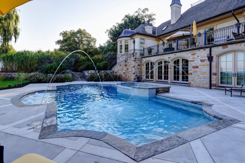 An elegant backyard with a curved swimming pool, stone house with balconies, hot tub, water feature, landscaped garden, and clear sky.