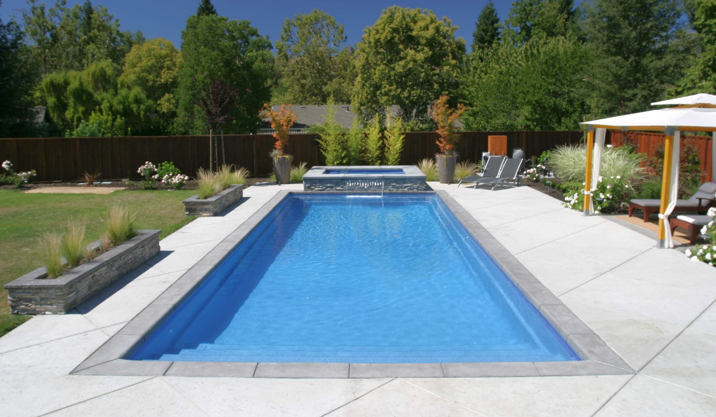 A luxurious backyard with a rectangular pool, sun loungers, landscaped garden, a pergola, and a well-manicured lawn on a sunny day.