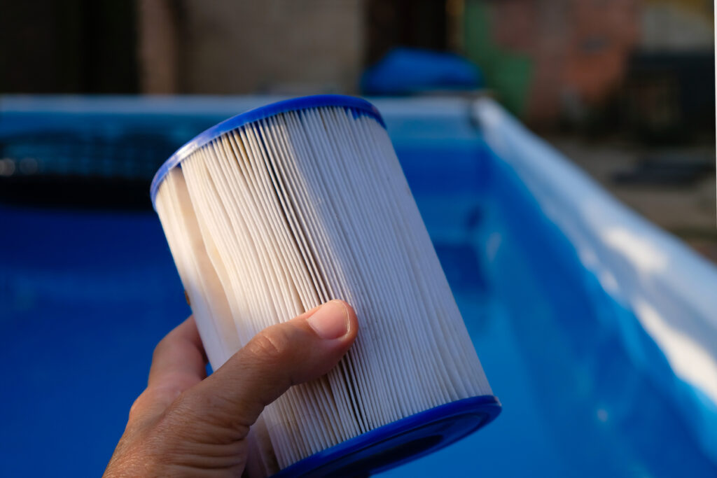 A person's hand is holding a large cylindrical white pool filter cartridge, with a blurred blue swimming pool in the background.
