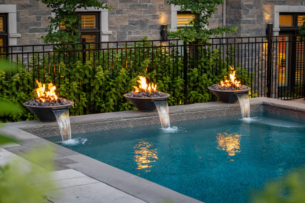 An outdoor swimming pool at dusk flanked by fire pits with flames, water features creating waterfalls, a fence, and a stone building in the background.