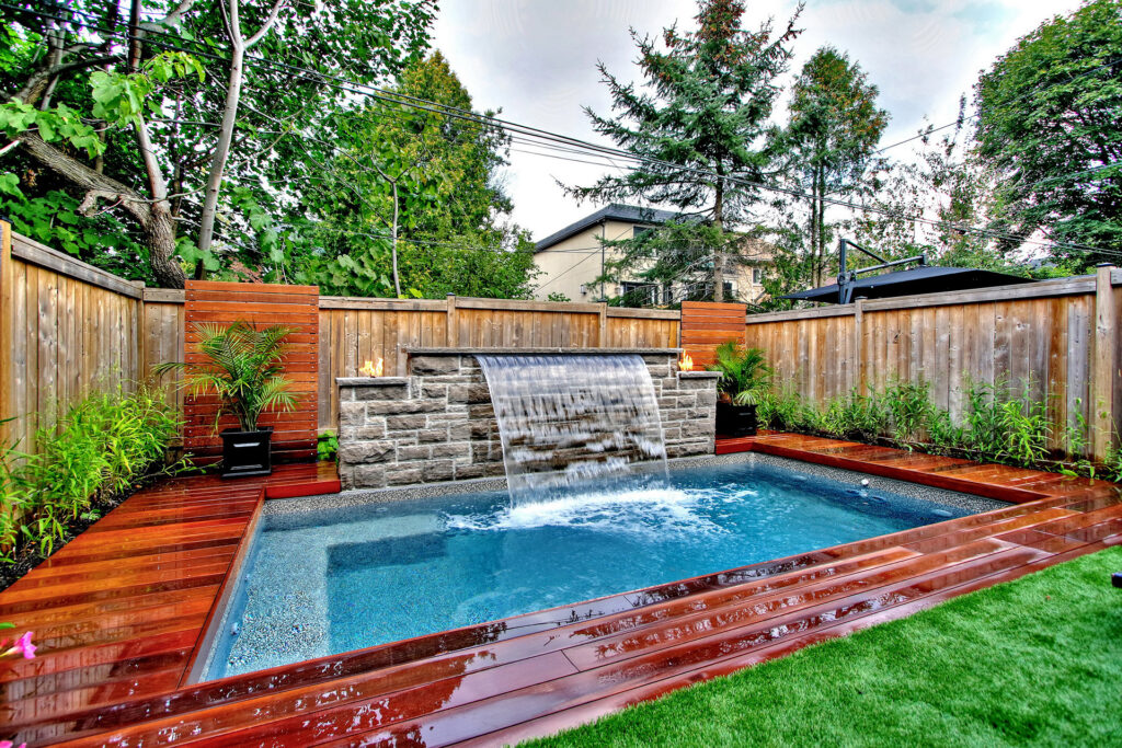 A landscaped backyard with a small rectangular pool, waterfall feature, wooden deck, and a fence surrounded by greenery under a cloudy sky.