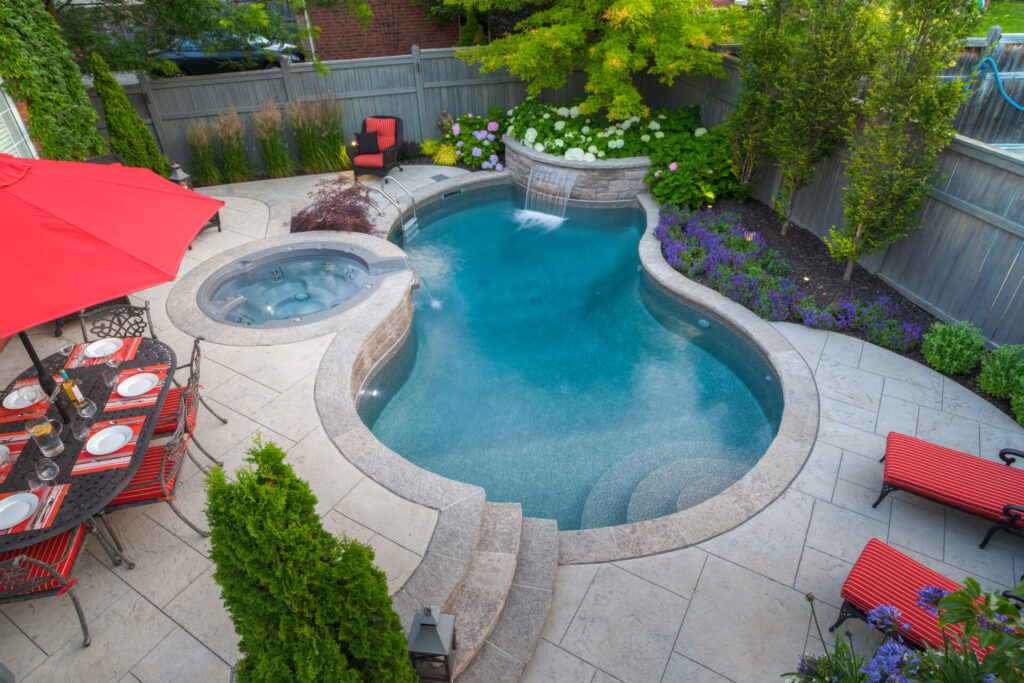 An elevated view of a landscaped backyard with a pool and hot tub, surrounded by flowering plants and arranged with outdoor dining and lounging furniture.
