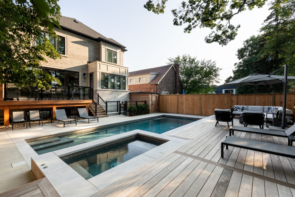 An elegant backyard with a swimming pool, hot tub, and wooden decking. Patio furniture is arranged neatly by the poolside, near a modern house.