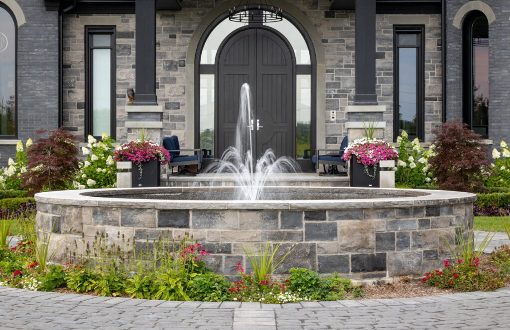 A landscaped entrance with a stone fountain, colorful flowers in planters, and a luxury house with dark stone detailing and a large door.