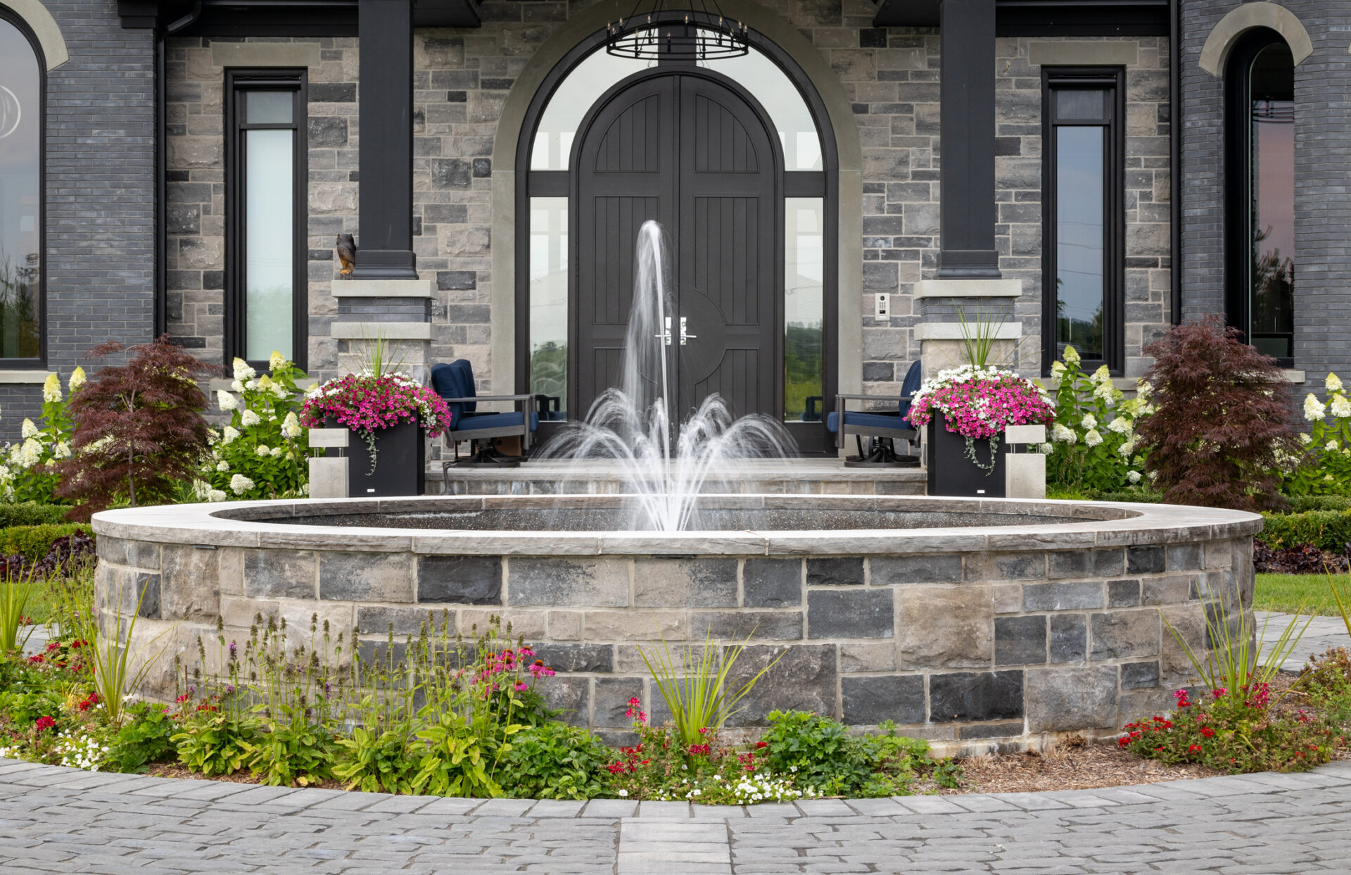 A landscaped entrance with a stone fountain, colorful flowers in planters, and a luxury house with dark stone detailing and a large door.