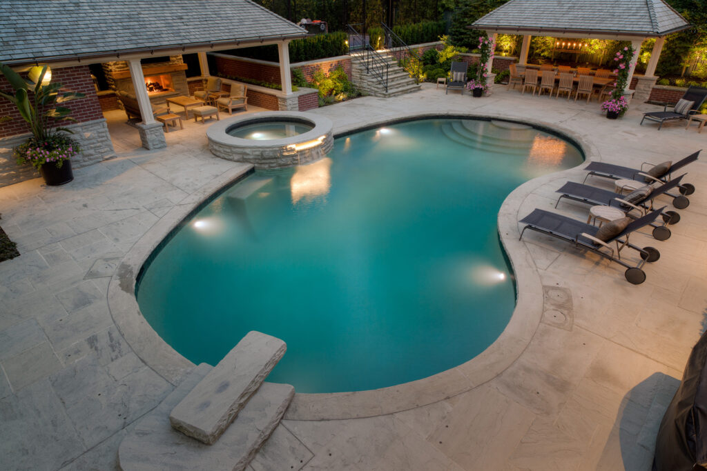 An elegant backyard with a curved swimming pool, hot tub, patio area, loungers, and covered outdoor seating with a fireplace at dusk.
