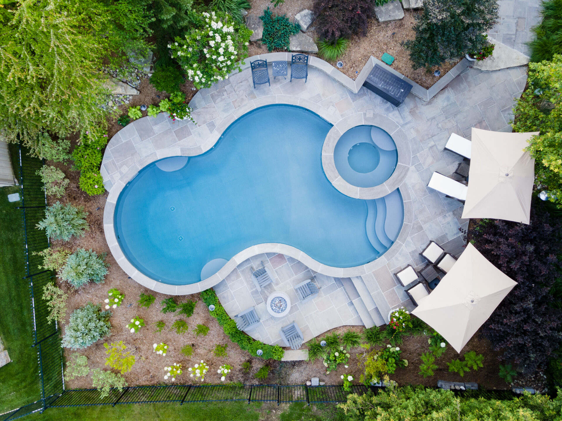Aerial view of a backyard with a uniquely shaped swimming pool, surrounded by patio furniture, landscaping, umbrellas, and a neatly manicured lawn.
