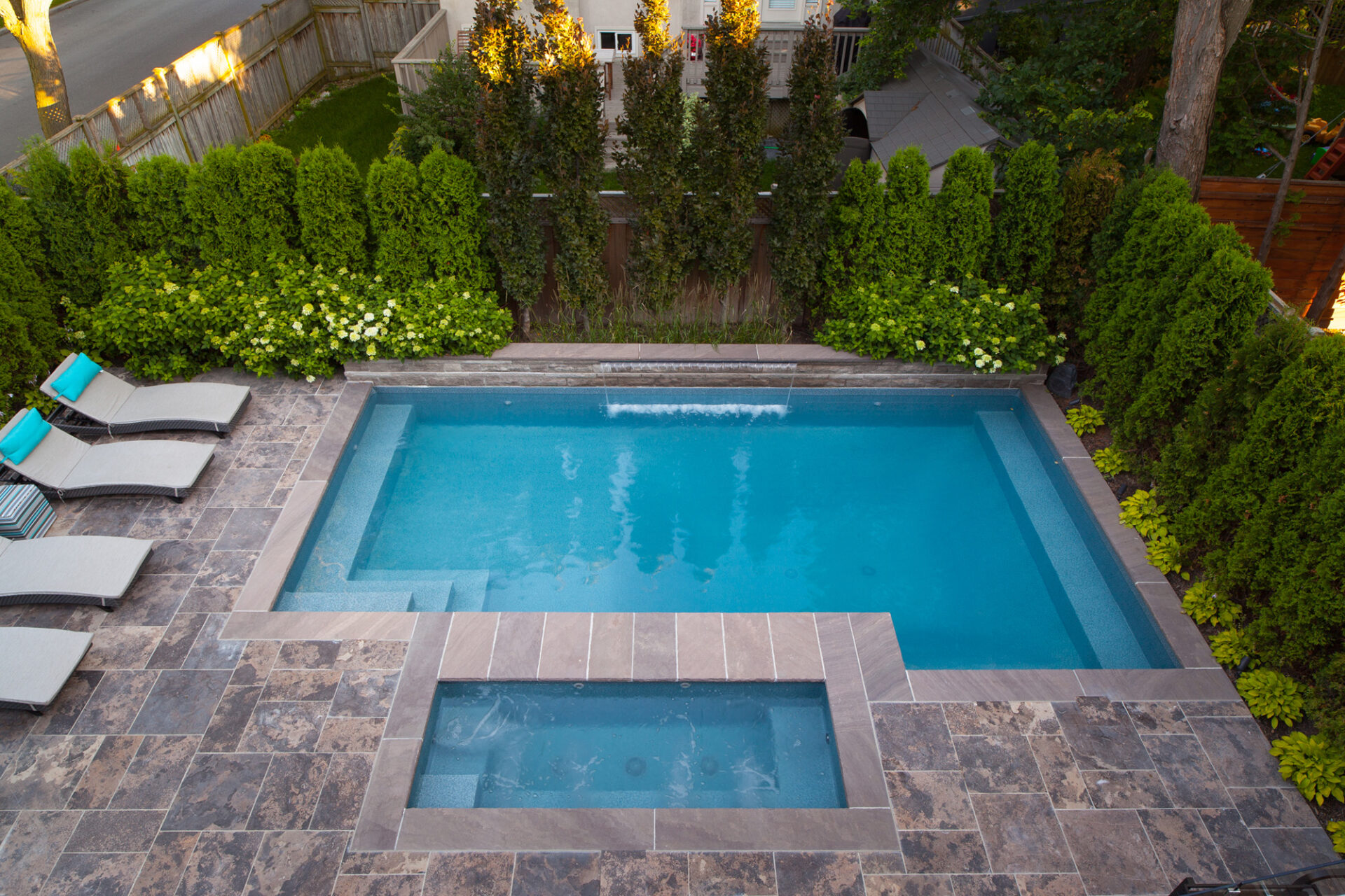 A serene backyard with a rectangular swimming pool and a hot tub, surrounded by lush greenery, stone tiles, and sun loungers with blue cushions.