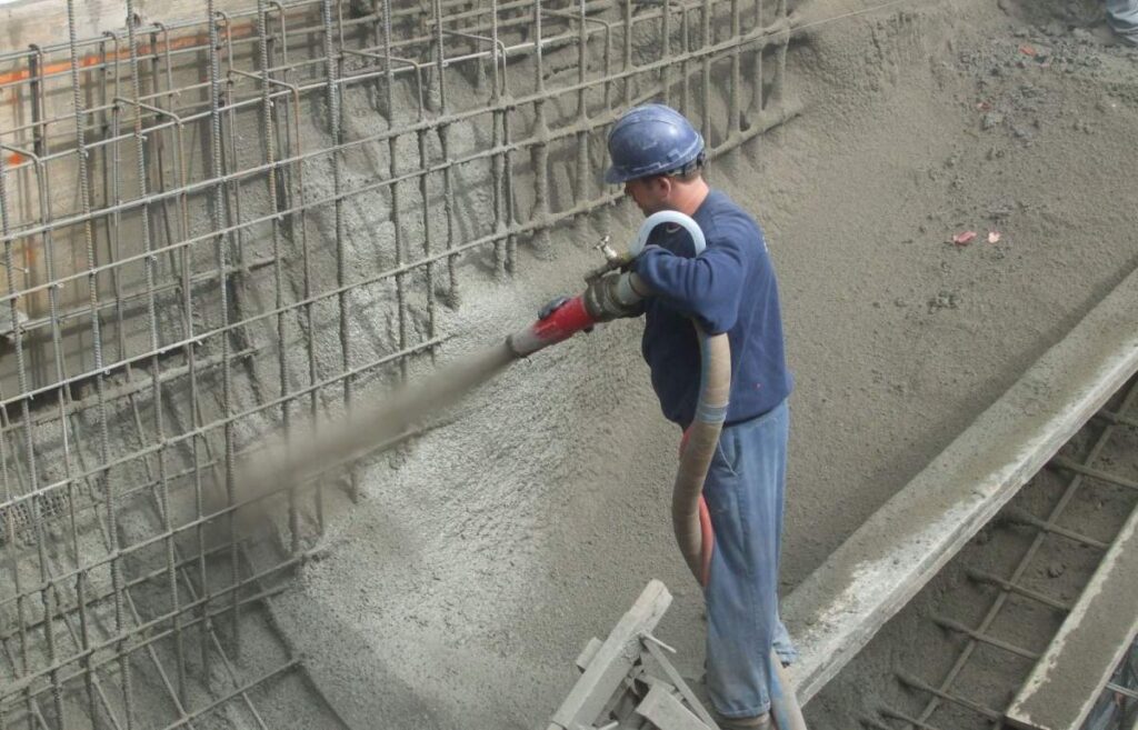 A person wearing a hard hat and overalls is spraying concrete onto a reinforced wall at a construction site, using a large hose.
