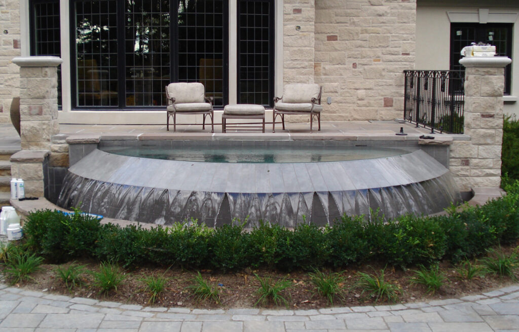 A semi-circular in-ground pool with a cover partially extended, flanked by two upholstered patio chairs, near a stone-faced building with large windows.