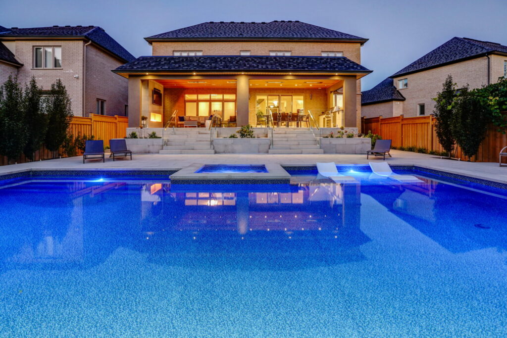 Luxurious backyard at dusk featuring a swimming pool with blue lights, cozy patio area, elegant house with illuminated interior, and a clear sky.