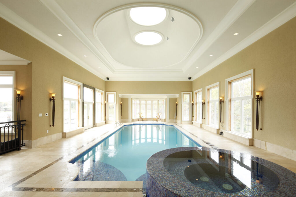 An indoor swimming pool with a vaulted ceiling, skylights, floor-to-ceiling windows, sconce lighting, and a hot tub tiled with a mosaic design.