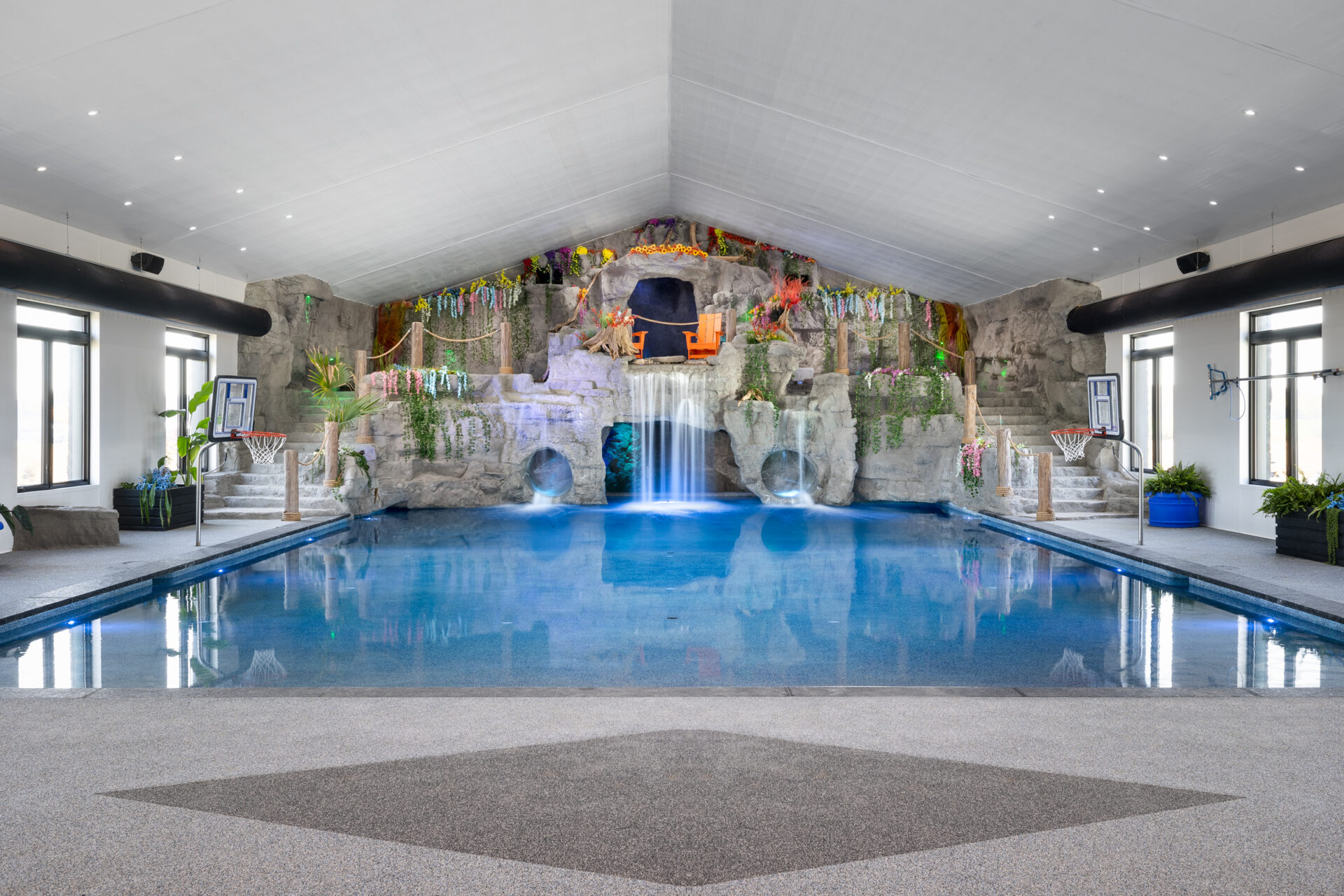 An indoor swimming pool with a decorative rocky waterfall and artificial foliage. Basketball hoops are installed on both sides with a symmetric design.