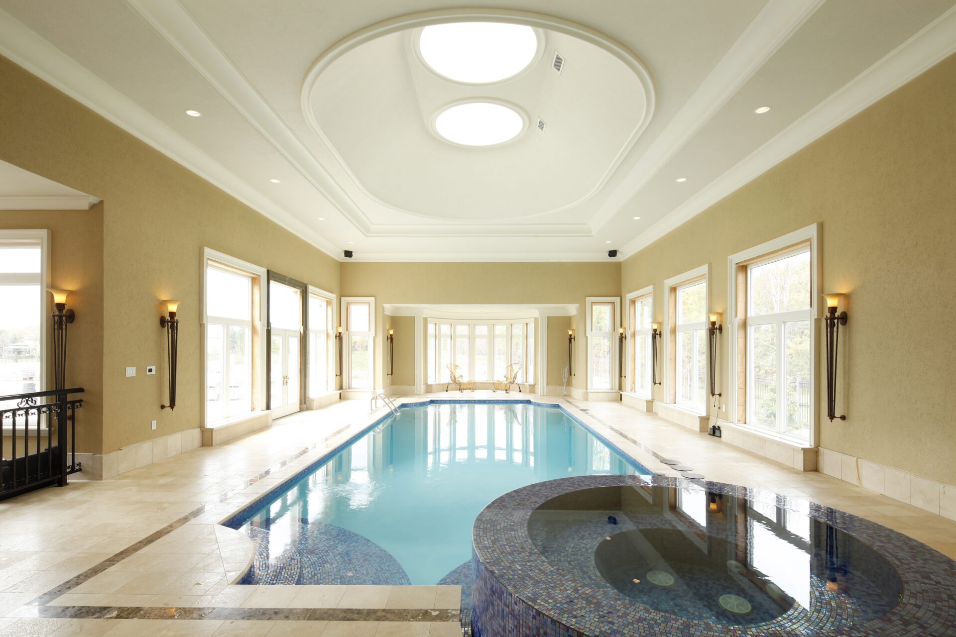 An elegant indoor swimming pool with arched ceilings, natural light from multiple windows, a small hot tub, and decorative wall sconces.
