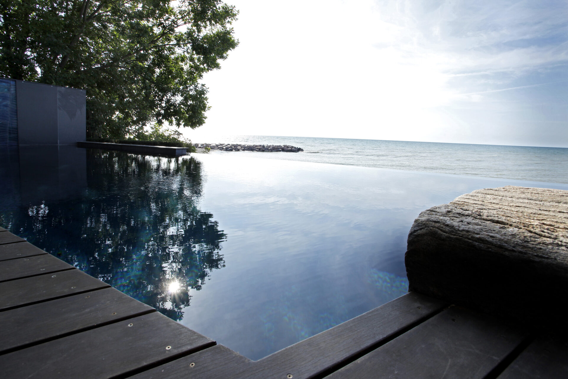 An infinity pool merges seamlessly with the ocean horizon, bordered by a wooden deck and a stone feature. Trees and sky reflect on the tranquil water.
