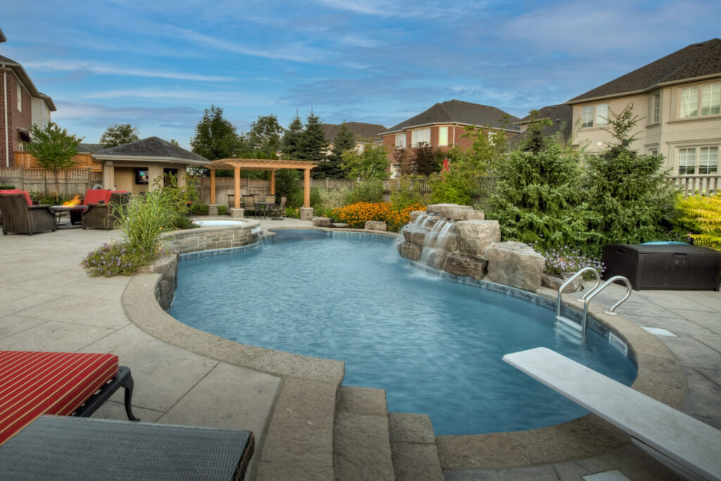A serene backyard with a curved swimming pool, waterfall feature, patio area with furniture, pergola, and landscaped garden against a backdrop of houses.