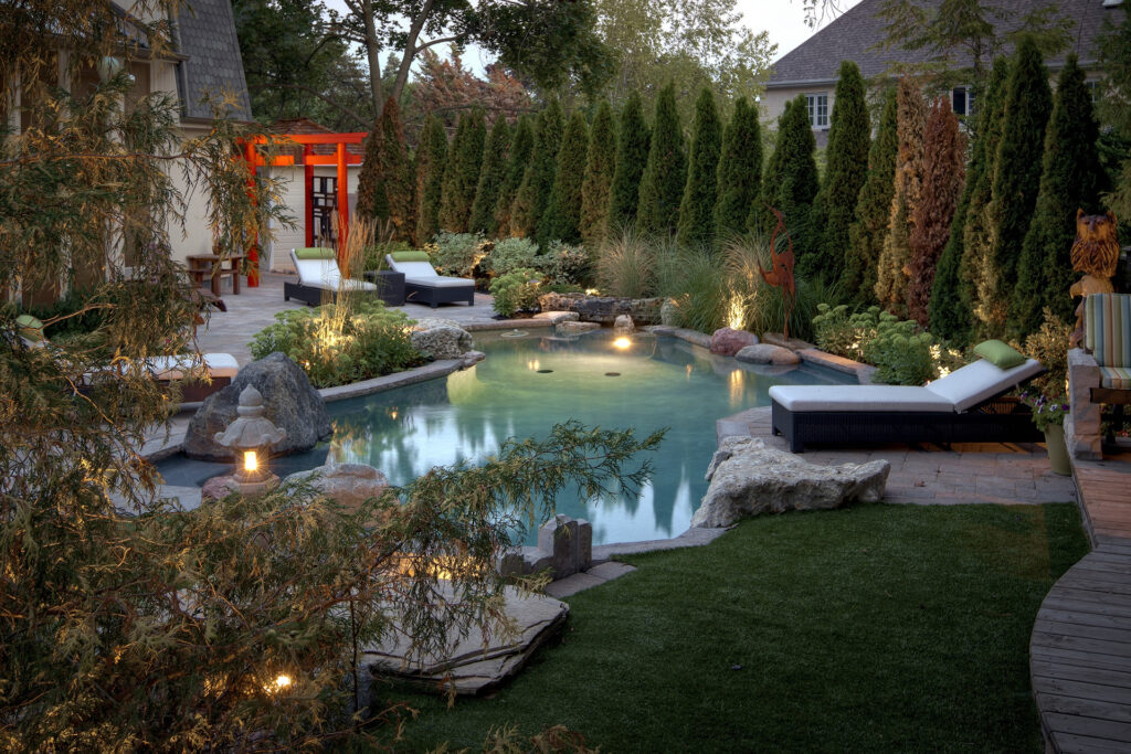 A backyard with a natural-style swimming pool, surrounded by lush landscaping, loungers, and illuminated by soft evening lights. A Torii gate stands prominently.
