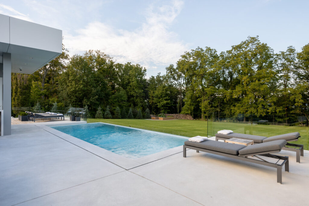 Modern backyard with a rectangular swimming pool, lounge chairs, neatly manicured lawn, trees, and part of a contemporary house visible. Clear skies above.